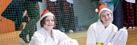 Dwoje dzieci w białych kimonach karate siedzi razem na hali sportowej w czerwonych czapkach Mikołaja, uśmiechnięte, w tle siatka i inni ćwiczący — klubowe spotkanie mikołajkowe