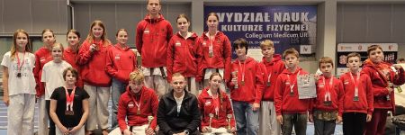 Drużyna sekcji karate Raion Fight Club w klubowych bluzach, część zawodników z medalami na szyi, wspólne zdjęcie po zawodach na hali sportowej